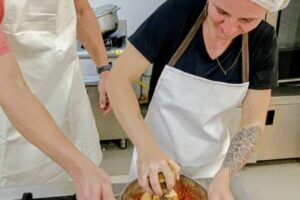 Two cooks wearing aprons and hair nets mix ingredients in a bowl over a stainless-steel kitchen counter.