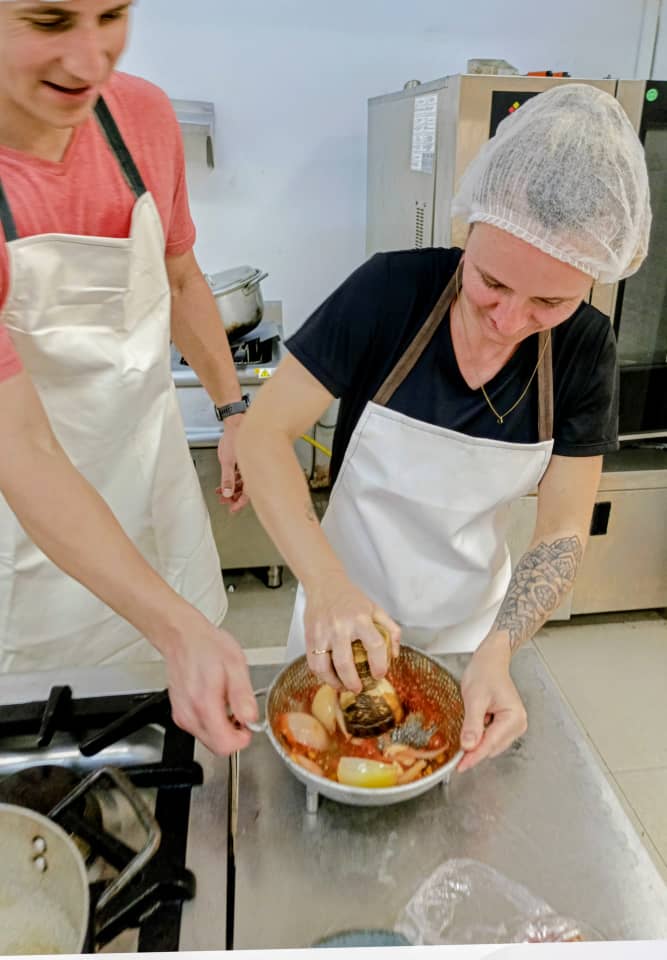 Two cooks wearing aprons and hair nets mix ingredients in a bowl over a stainless-steel kitchen counter.