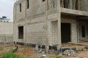 Two-story unfinished concrete block house under construction with exposed rebar and wooden formwork on the upper level, debris in the foreground.