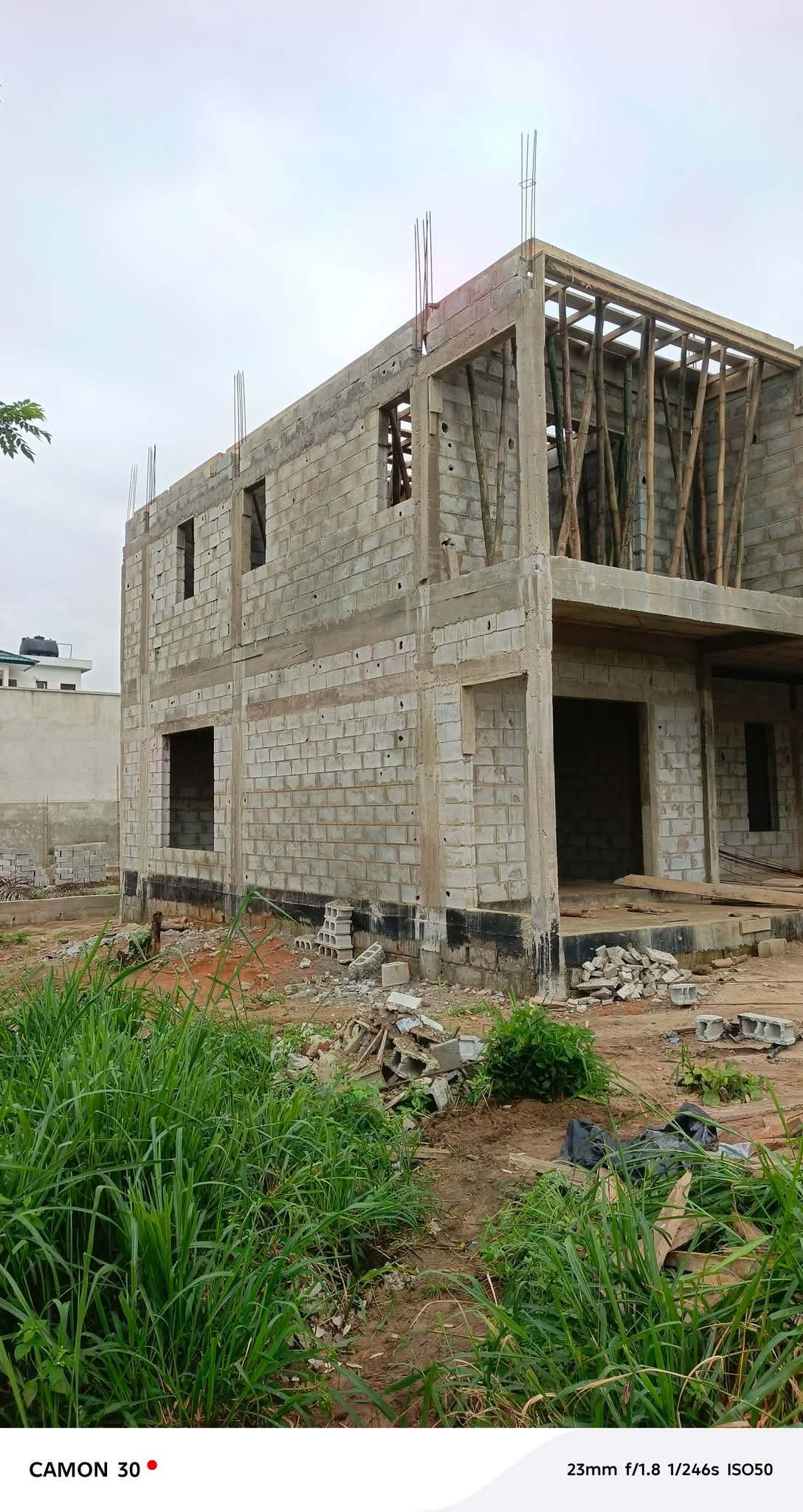 Two-story unfinished concrete block house under construction with exposed rebar and wooden formwork on the upper level, debris in the foreground.